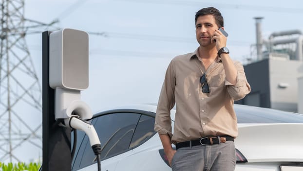 Man talking on the phone while recharge EV car battery at charging station connected to power grid tower electrical as electrical industry for eco friendly car utilization. Panorama Expedient