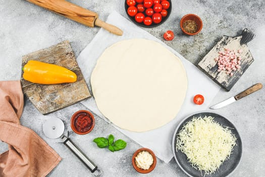 Pizza ingredients: cherry tomato, pepper, cheese, ham, basil with dough rolled out in a circle and a wooden chip lie on the table with copy space, top view close-up. Concept of cooking pizza.