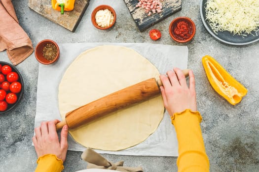 Hands of a caucasian teenager in an apron roll out pizza dough with a rolling pin on a table with ingredients, a knife and cutting boards, close-up top view. Pizza cooking concept.