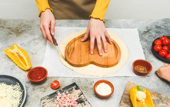Hands of a caucasian teenage girl in an apron cut out the dough in the shape of a heart for pizza for valentine's day with ingredients on the table, close-up side view.