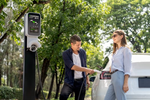 Lovely young couple wearing sun glasses recharging battery for electric car during road trip travel EV car in natural forest or national park. Eco friendly travel during vacation and holiday. Exalt