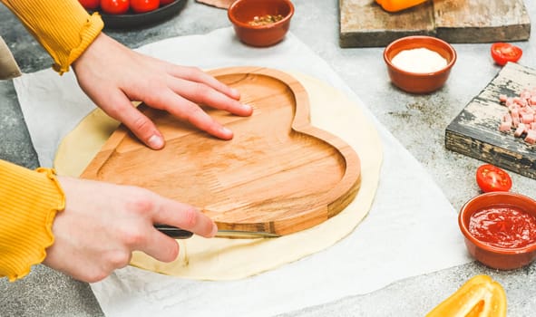 Hands of a caucasian teenage girl in an apron cut out the dough in the shape of a heart for pizza for valentine's day with ingredients on the table, close-up side view.Concept of cooking pizza valentine day