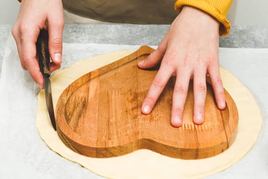Hands of caucasian teenage girl in apron cut out of heart shaped dough for valentine's day pizza on table, close-up side view. Concept of cooking pizza for valentine's day.