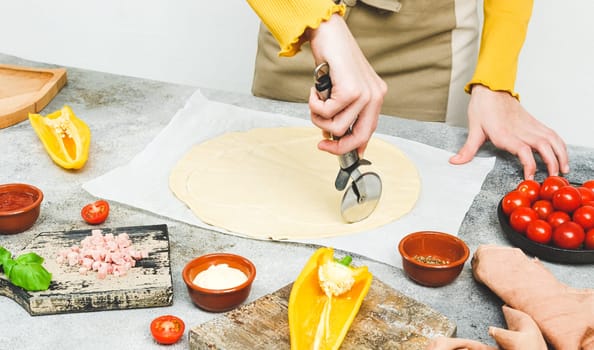 Hands of a caucasian teenage girl in an apron cut out the pizza dough in the shape of a heart with a scabbard for valentine's day with ingredients on the table, top view close-up.