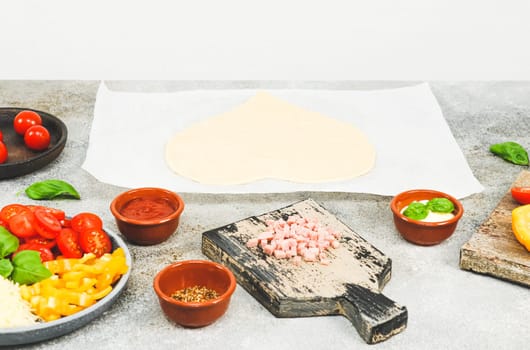 Heart shaped dough on baking paper with wooden boards and pizza ingredients lie on a light gray table, close-up side view.