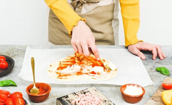 Hands of a Caucasian teenage girl smear a spoonful of pizza sauce on a heart-shaped dough on baking paper with wooden boards and pizza ingredients lie on a light gray table, side view close-up.