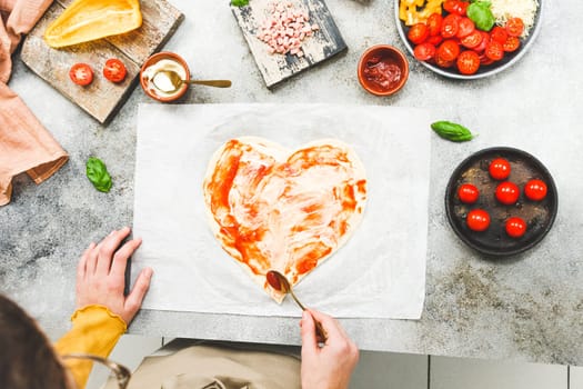 Hands of a caucasian teenage girl in an apron smear tomato sauce on a heart-shaped pizza dough for valentine's day with ingredients on the table, close-up side view. Valentine's day pizza cooking concept.