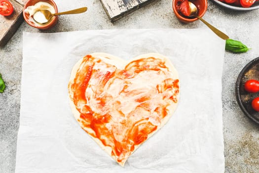 Carved from heart pizza dough on baking paper with sauces and ingredients on gray cement table, top view close-up. Concept of cooking pizza.