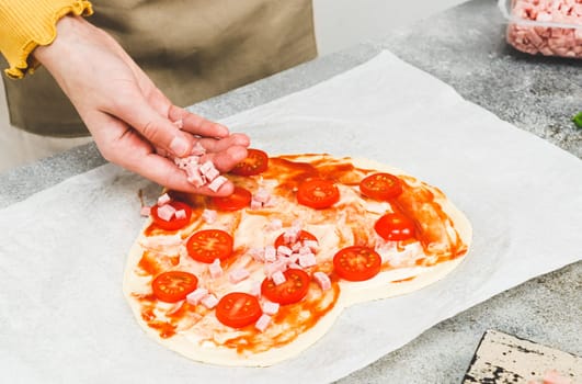 Hands of caucasian teenage girl lay down sliced cherry tomato on heart-shaped pizza dough for valentine's day with ingredients on the table, close-up side view. Valentine's day pizza making concept.