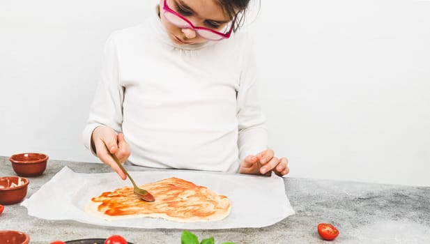 Caucasian girls in a white turtleneck smear a spoon with tomato sauce on a heart-shaped pizza dough for valentine's day with ingredients on the table, close-up side view. Valentine's day pizza cooking concept.