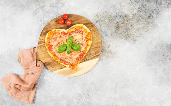 Hot pizza heart on a round wooden cutting board with cherry tomatoes and a kitchen napkin lie on the left on a gray cement table with copy space on the right, top view close-up. Fast food concept.