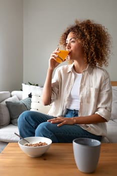 Vertical portrait of young multiracial woman drinking orange juice for breakfast sitting on sofa at home living room looking away pensive.Lifestyle concept.