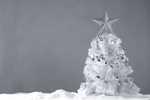 White christmas tree with silver decorations and gifts on snow on gray background