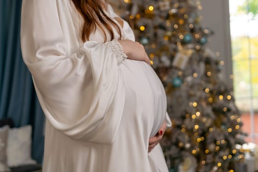 Pregnant woman holding a belly against Christmas tree with lights. family holiday concept