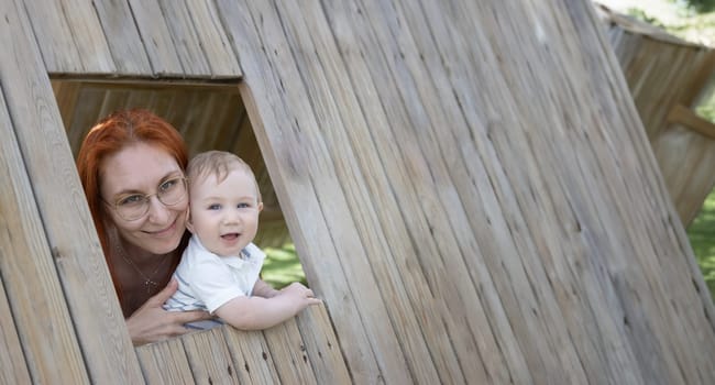 Smiling baby and his mother sits in a wooden house on the playground - looking in the camera. Mid shot