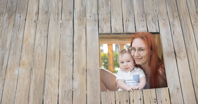 Cute baby and his mother sits in a wooden house on the playground - looking in the camera. Mid shot