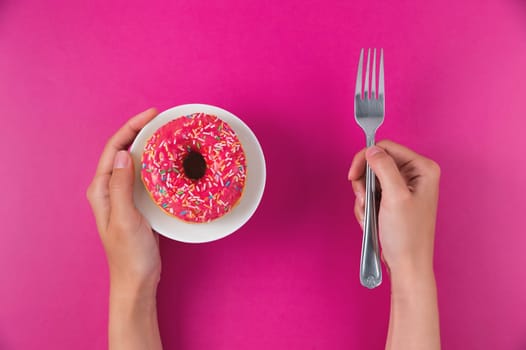 Female hands hold a donut on a plate and a fork on a pink background. Top view, flat lay. Sweet, diet concept. Losing weight after the holidays.