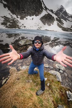 A Caucasian male mountain traveler in sunglasses and trekking clothes expressively invites you to the mountains against the backdrop of snow-covered rocks and an alpine lake. Welcome to tourism.