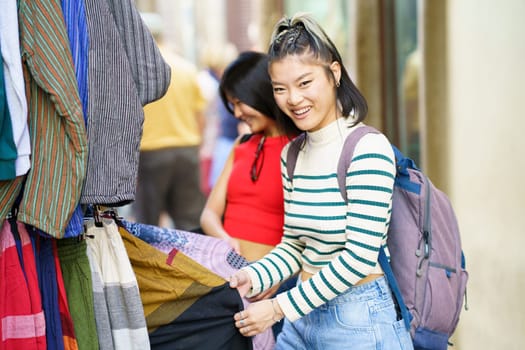 Positive young Asian female, in casual clothes with backpack smiling and looking at camera while shopping clothes with friend on street market in city during vacation