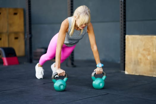 Strong fit young woman in activewear doing push ups exercise with heavy kettlebells during fitness session at modern gym on floor against blurred background