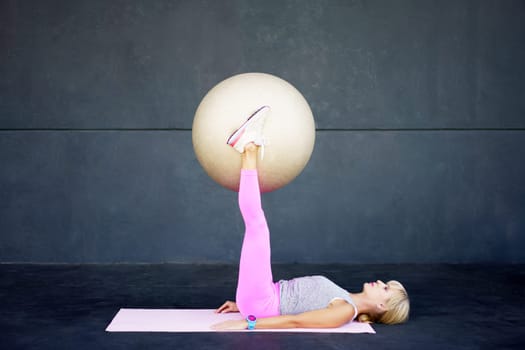 Side view of flexible young fit female, lying on mat and doing pilates exercise with fitness ball while training in gym against gray wall during workout