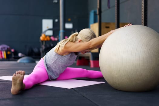 Side view of anonymous barefoot female athlete in sportswear, looking down while sitting with legs apart on mat with hands on fit ball and doing stretching exercise in gym in daylight