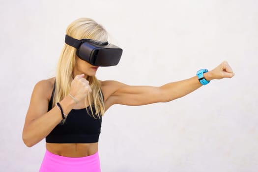 Fit female with long blond hair wearing VR goggles and boxing with hands in air while having virtual reality experience in sport against white background