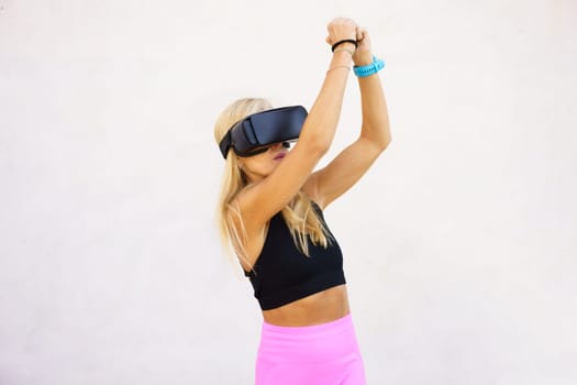 Young fit woman in VR goggles playing videogame while raising arms over head with invisible object against white background