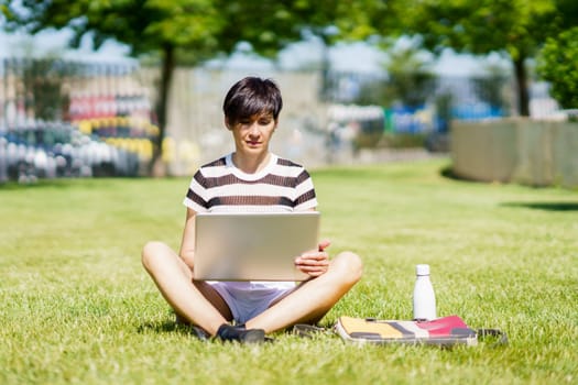 Full body of focused female freelancer in casual clothes sitting on grass and browsing laptop while working on remote project in park