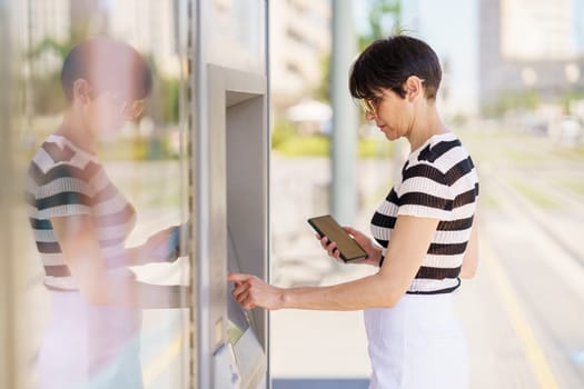 Concentrated adult female in casual outfit and sunglasses standing with smartphone and buying ticket on terminal on street