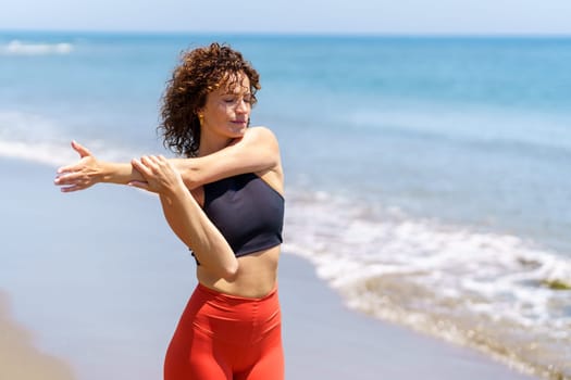 Pensive female athlete with closed eyes and curly hair stretching arm during workout on sunny day at beach