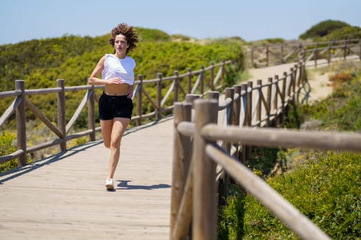Full body of young active female in sportswear jogging on wooden walkway during fitness workout on sunny summer day