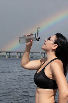 A beautiful and slender girl in sunglasses pours water over herself from a sports bottle after a fitness workout on the street in the summer, against the sky with a rainbow. Sport concept. Close-up.