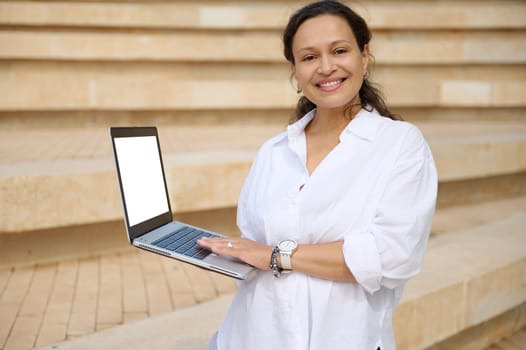 Multi-ethnic pleasant smiling businesswoman broadly smiling looking at camera, holding laptop with white mockup digital screen. Copy advertising space. People. IT technology. Online Business. Startup