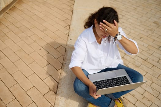 View from above of a businesswoman feeling upset, touching forehead while failing business plan or losing tender, sitting outdoors on steps and online working on laptop. People. Brainstorming. Career