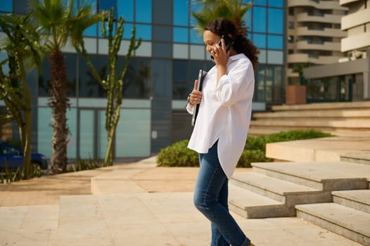 Young female manager in casual attire, talking on mobile phone while walking in the city. Confident successful business woman or entrepreneur freelancer, working outdoors, cityscape in the background.