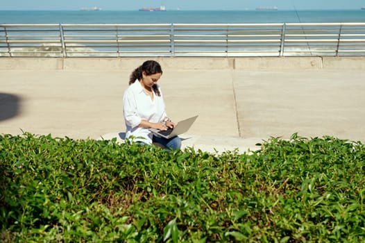 Authentic confident young multi ethnic businesswoman working on laptop outdoor, sitting on bench against Atlantic ocean background. Green shrubs on the foreground . Remote work and online job concept