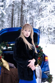 Woman in winter snowy forest in blue dress next to blue car decorated with Christmas decor. Christmas and winter holidays concept