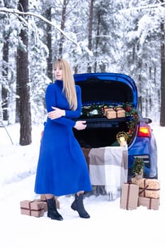 Woman in winter snowy forest in blue dress next to blue car decorated with Christmas decor. Christmas and winter holidays concept