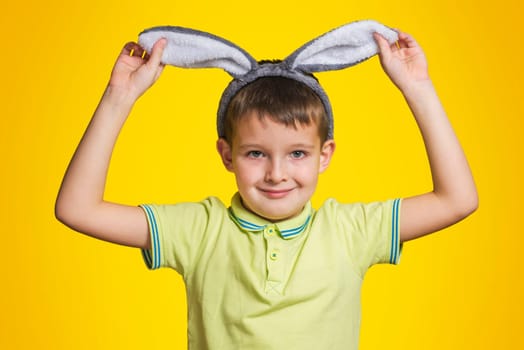 Little boy in t-shirt is touching his bunny ears and smiling while standing yellow background. Cute little child wearing bunny ears on Easter day.