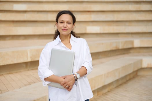 Confident portrait of a beautiful young adult business woman holding laptop, looking confidently at camera standing against steps background. People. Professional occupation. Career and recruitment