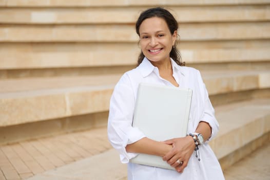 Confident successful charming young adult business woman holding laptop,smiling looking confidently at camera standing against steps background. People. Professional occupation. Career and recruitment