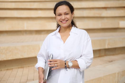 Portrait of multi ethnic pretty woman holding laptop, smiling looking at camera, standing against beige marble city steps backdrop, dressed in white casual shirt. Business people. Remote job. Career