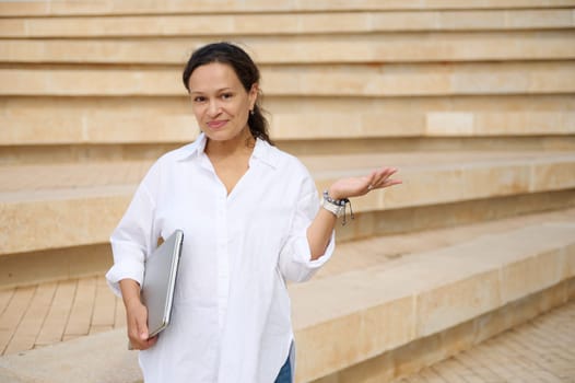 Confident authentic young mixed race woman in white casual shirt, holding laptop and imaginary copy space on her hand palm up, smiling looking at camera, standing against beige marble steps background