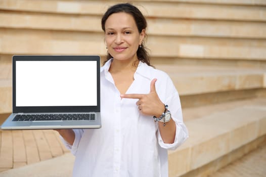 Beautiful multi-ethnic young woman in casual white shirt, pointing index finger at white mockup digital screen with copy advertising space, smiling looking at camera standing against steps background