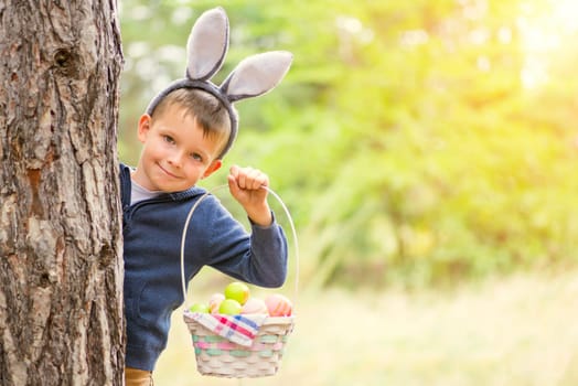 Happy child looks out of a tree holding basket full of colorful easter eggs after egg hunt at spring time. Happy Easter day.