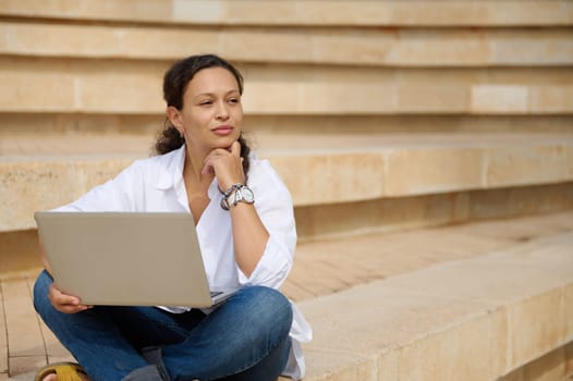 Young thoughtful confident female freelancer entrepreneur, sales manager using laptop outdoors, looking into the distance, thinking on new strategy of business development, sitting on steps outdoors