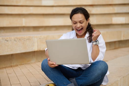 Young woman student clenching fist, raising arm up, smiling, expressing happiness positive emotions while passing university exams and goals achievement, sitting outdoors on steps with laptop on knees
