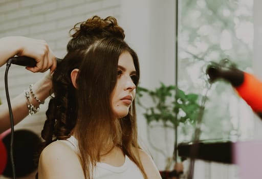 Portrait of a young beautiful brunette Caucasian girl sitting in a hairdresser's shop, where the master is curling her long hair on a flat iron, close-up view from below.