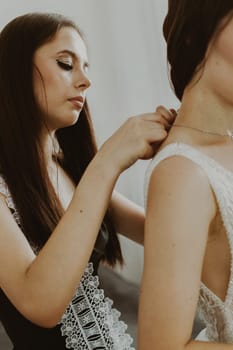 Beautiful young brunette bridesmaid fastens the clasp of a necklace around the neck of the bride standing in a room by the window, close-up side view.